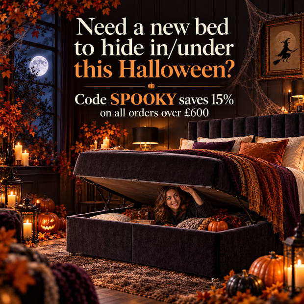 A dark upholstered storage bed with the lift-up mattress platform open, showing a woman smiling underneath in a cozy Halloween-themed room decorated with pumpkins, candles, and autumn leaves. Promotional text overlays read: 'Need a new bed to hide in/under this Halloween? Code SPOOKY saves 15% on all orders over £600'.
