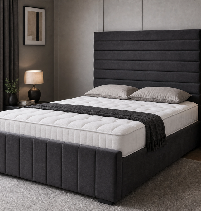 The Belmont Divan Ottoman Bed in charcoal grey velvet with horizontal pleated headboard and vertical panel footboard, shown closed with mattress and pillows in a modern bedroom setting, available in all UK sizes